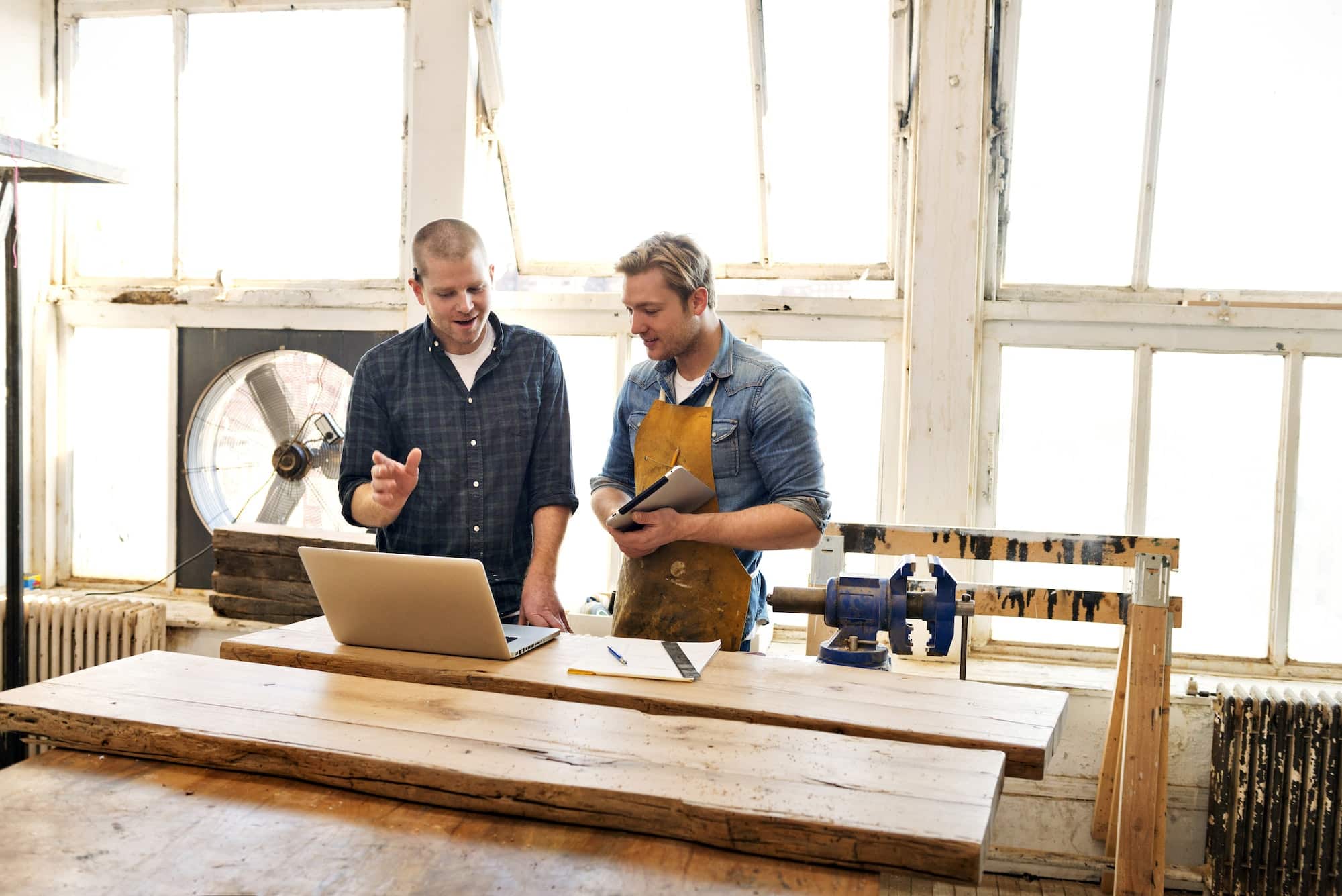 Male Carpenters Looking At Laptop And Discussing In Workshop Male Carpenters Looking At Laptop And Discussing In Workshop